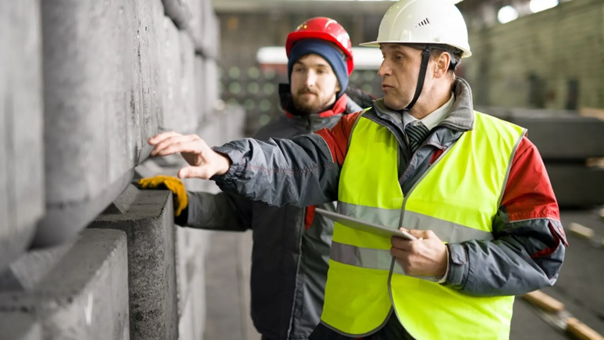 portret van volwassen werknemer met warme jas en veiligheidshelm met behulp van digitale tablet terwijl hij de productie van betonblokken bespreekt met stagiair in werkplaats