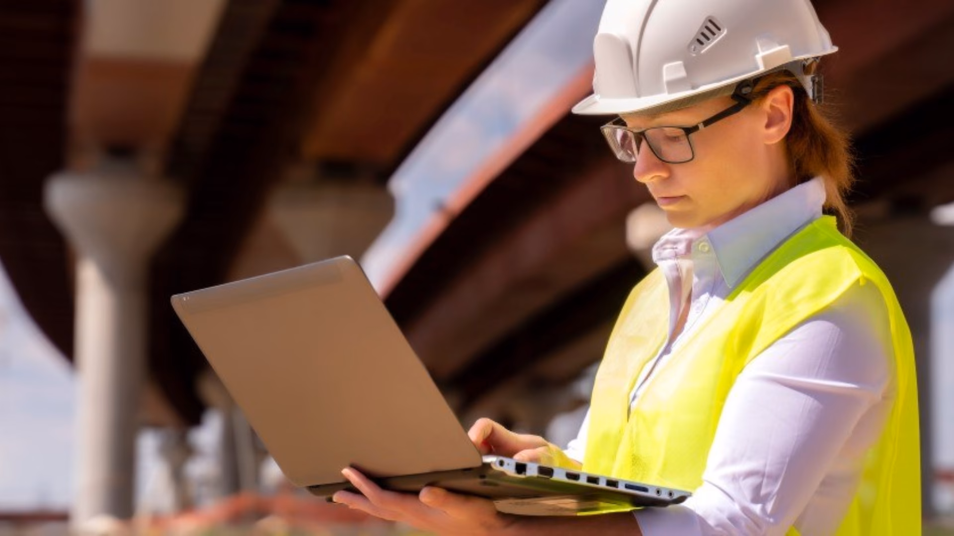 Een werkvoorbereider in een geel hesje en met een witte helm werkt onder een brug op een laptop.