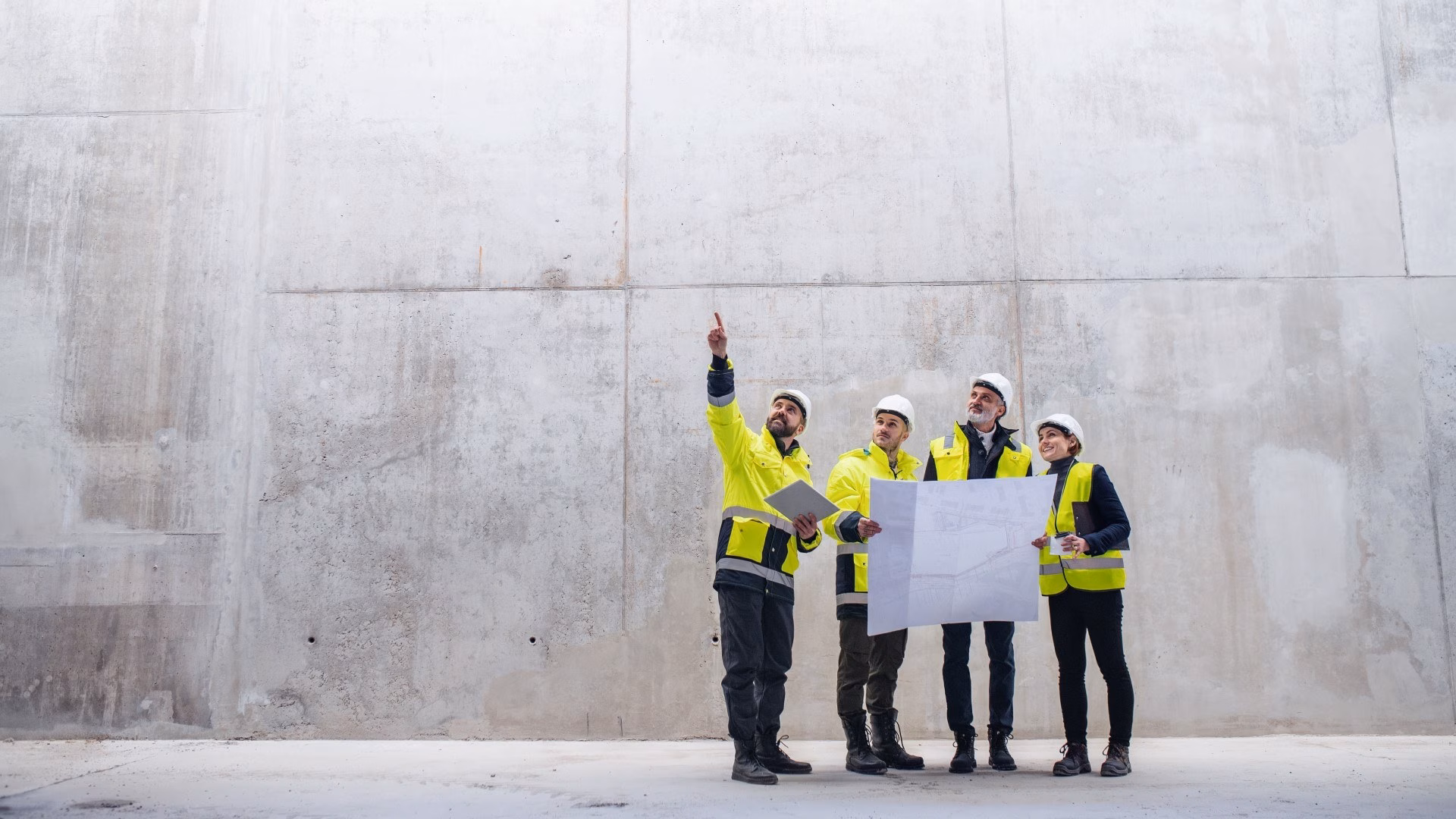 Een groep ingenieurs in gele hesjes en witte helmen staat voor een betonnen muur en bekijkt een bouwtekening