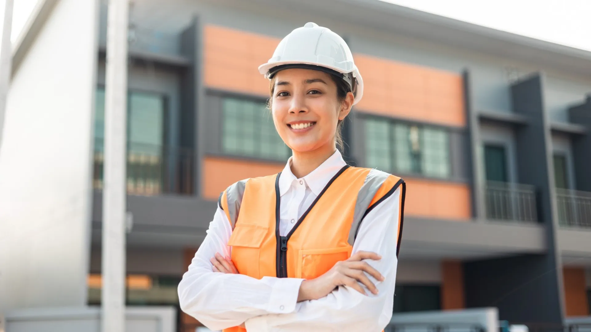 Een glimlachende ingenieur in een oranje hesje en een witte helm staat voor een moderne woning