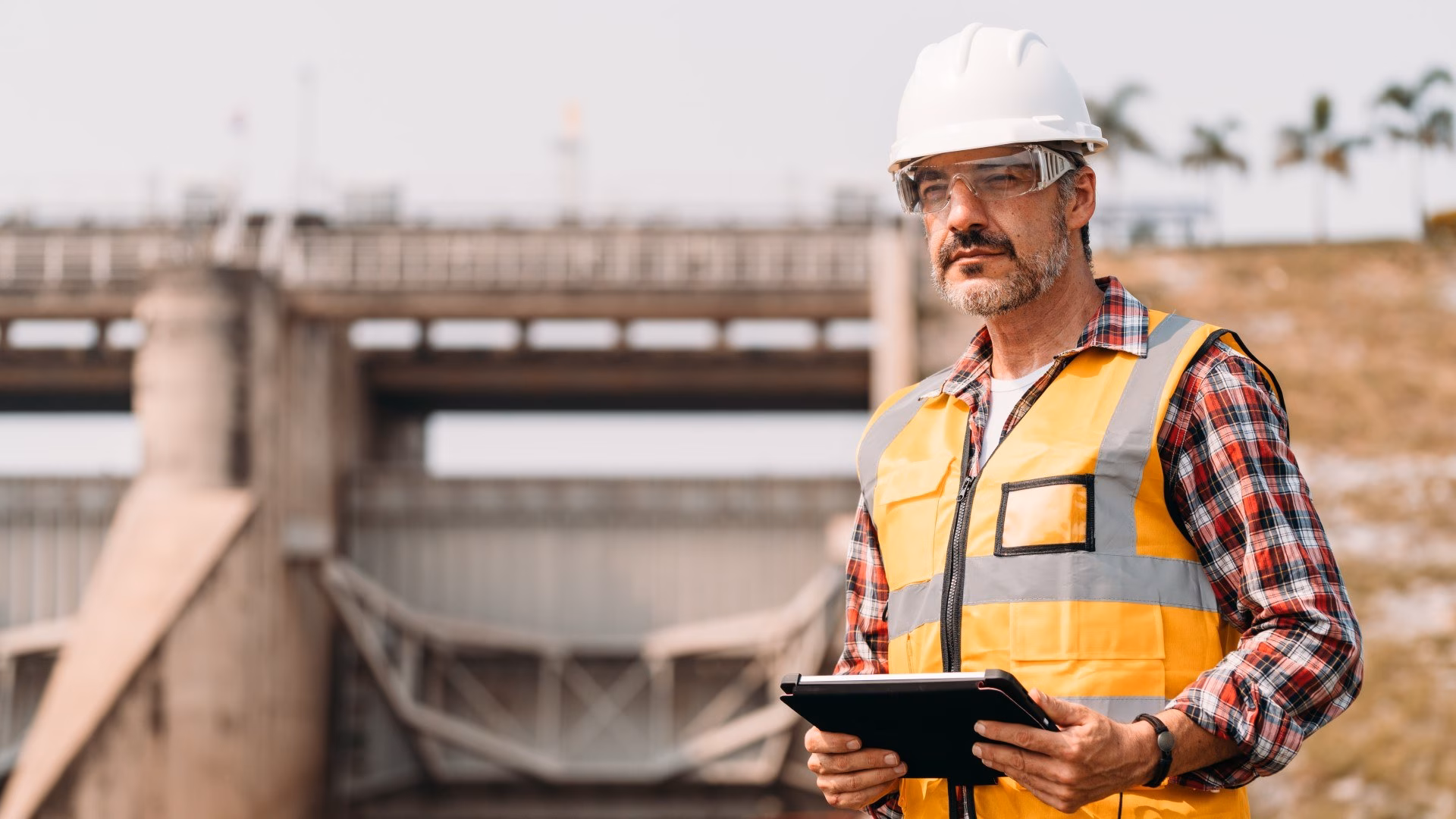 Een ingenieur met een witte helm, veiligheidsbril en oranje hesje houdt een tablet vast met een betonnen waterwerk op de achtergrond