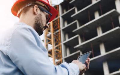 Een ingenieur in een oranje helm tikt op zijn telefoon terwijl hij voor een hoog gebouw in aanbouw staat.