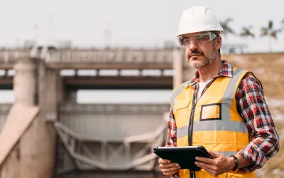 Een ingenieur met een witte helm, veiligheidsbril en oranje hesje houdt een tablet vast met een betonnen waterwerk op de achtergrond