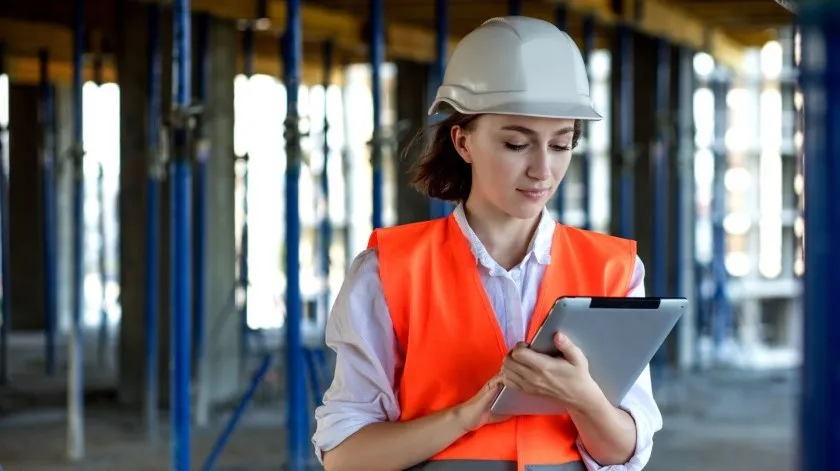 Een ingenieur op een bouwplaats in een oranje hesje en met een witte helm noteert bevindingen op een tablet.