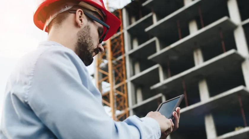 Een ingenieur in een oranje helm tikt op zijn telefoon terwijl hij voor een hoog gebouw in aanbouw staat.