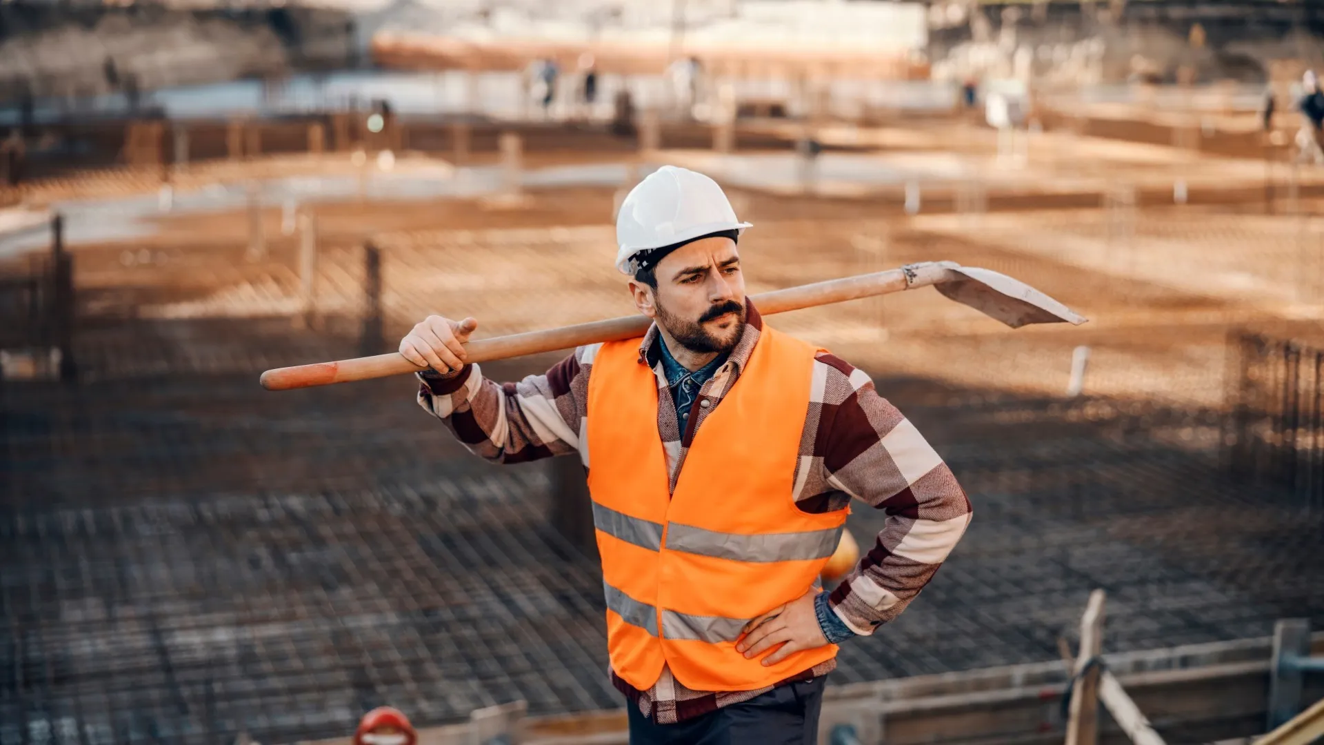 Een bouwplaatsmedewerker staat met een schop over zijn schouder op een bouwplaats met veel wapening