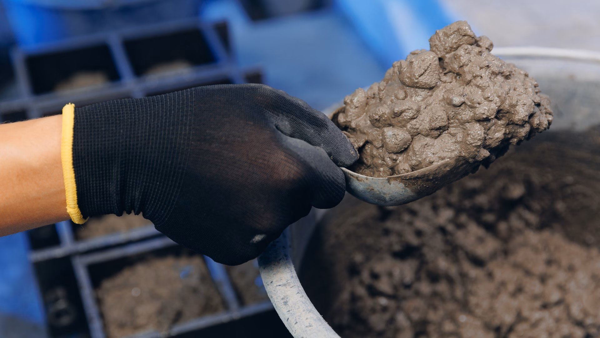 Een medewerker in het lab met een zwarte handschoen aan schept betonspecie uit een emmer om betonkubussen te maken om te testen.