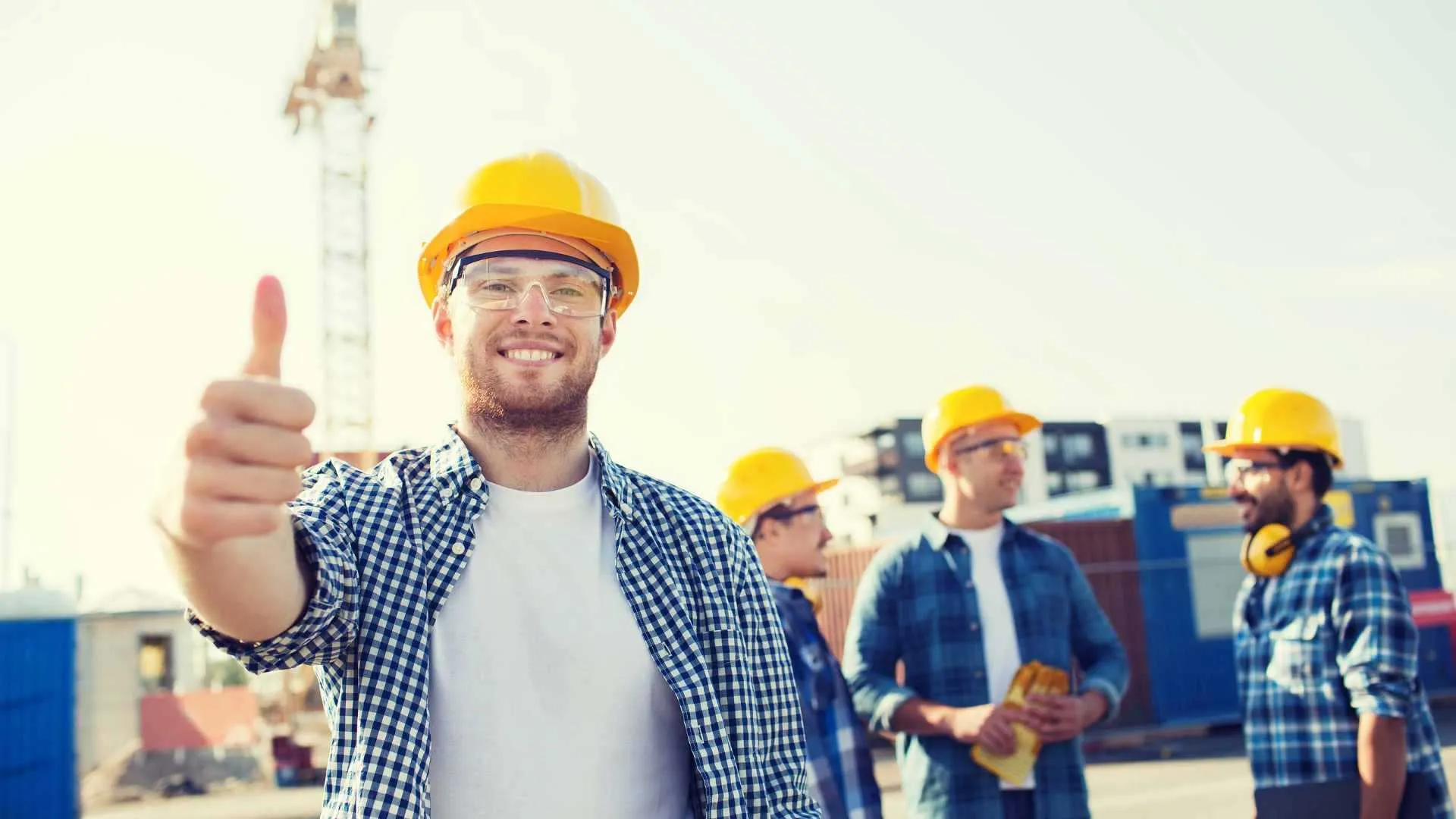 Een bouwvakker met gele helm en veiligheidsbril staat met zijn duim omhoog op een bouwplaats met een bouwkraan en drie collega's achter zich.
