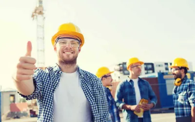 Een bouwvakker met gele helm en veiligheidsbril staat met zijn duim omhoog op een bouwplaats met een bouwkraan en drie collega's achter zich.